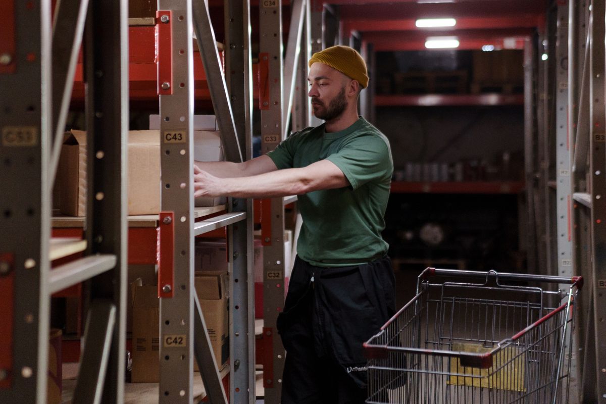 A man stocking boxes onto a warehouse shelf beside a shopping cart