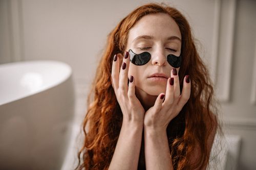 Free Woman with red hair enjoying a skincare routine with under-eye masks indoors. Stock Photo