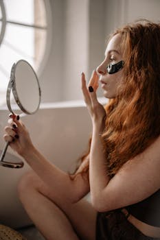 Young woman with red hair applying an eye mask while sitting by a window.