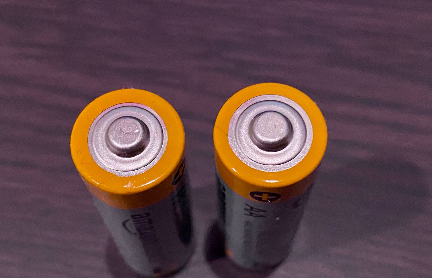 Top view of two AA batteries with yellow tops on a dark surface.