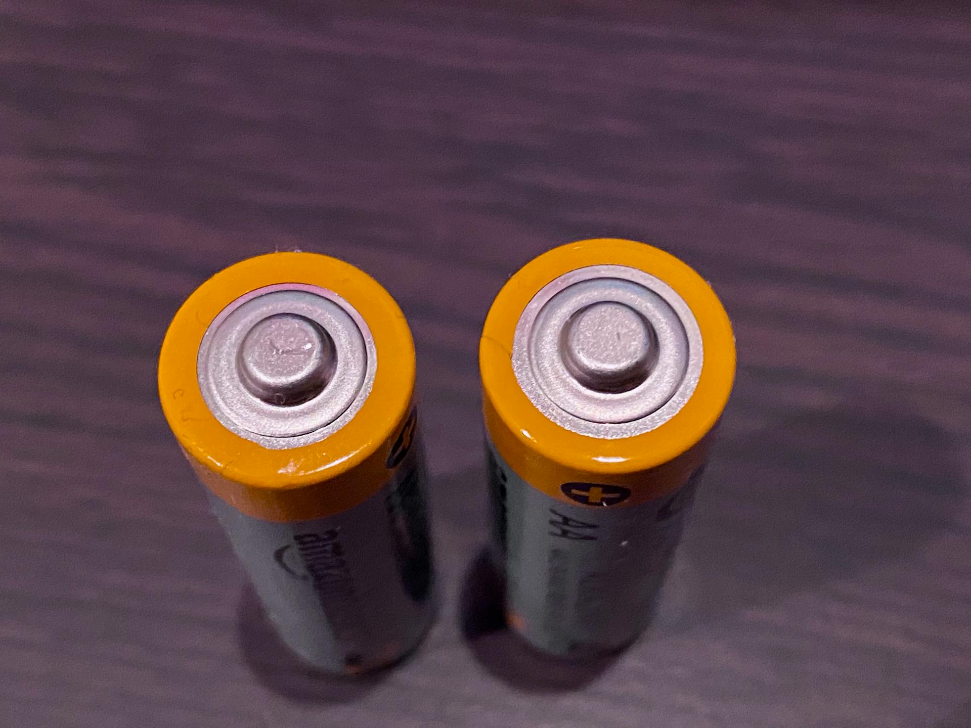 Top view of two AA batteries with yellow tops on a dark surface.