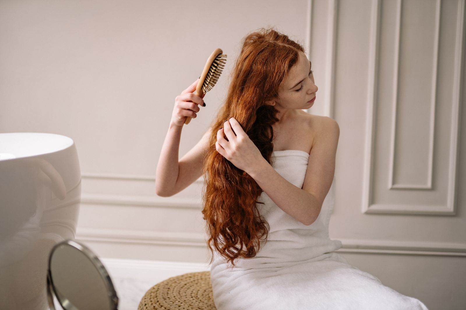 Woman with long wavy red hair, wrapped in a white towel, brushing her hair in a bright bathroom