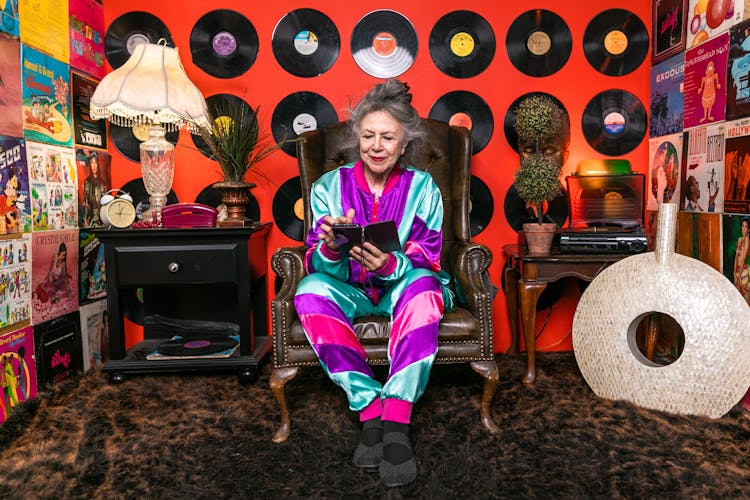 Elderly Woman In Purple And Pink Outfit Sitting On Brown Leather Armchair