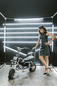 Stylish woman in black leather posing elegantly with a motorbike indoors.
