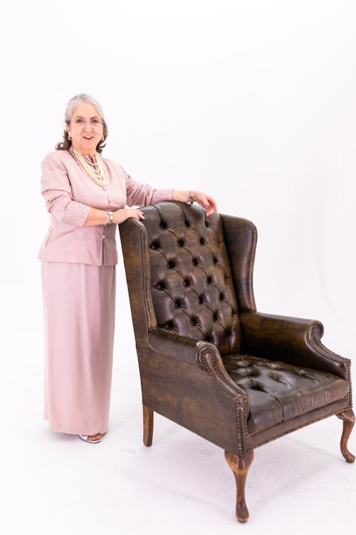 Woman in Pink Long Sleeve Dress Standing Beside Brown Wooden Armchair