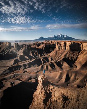 A lone person stands atop a cliff overlooking dramatic canyonland formations under a vibrant sky.