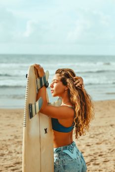 Woman with surfboard stands on a sunny beach, looking at the ocean. Beautiful summer day.