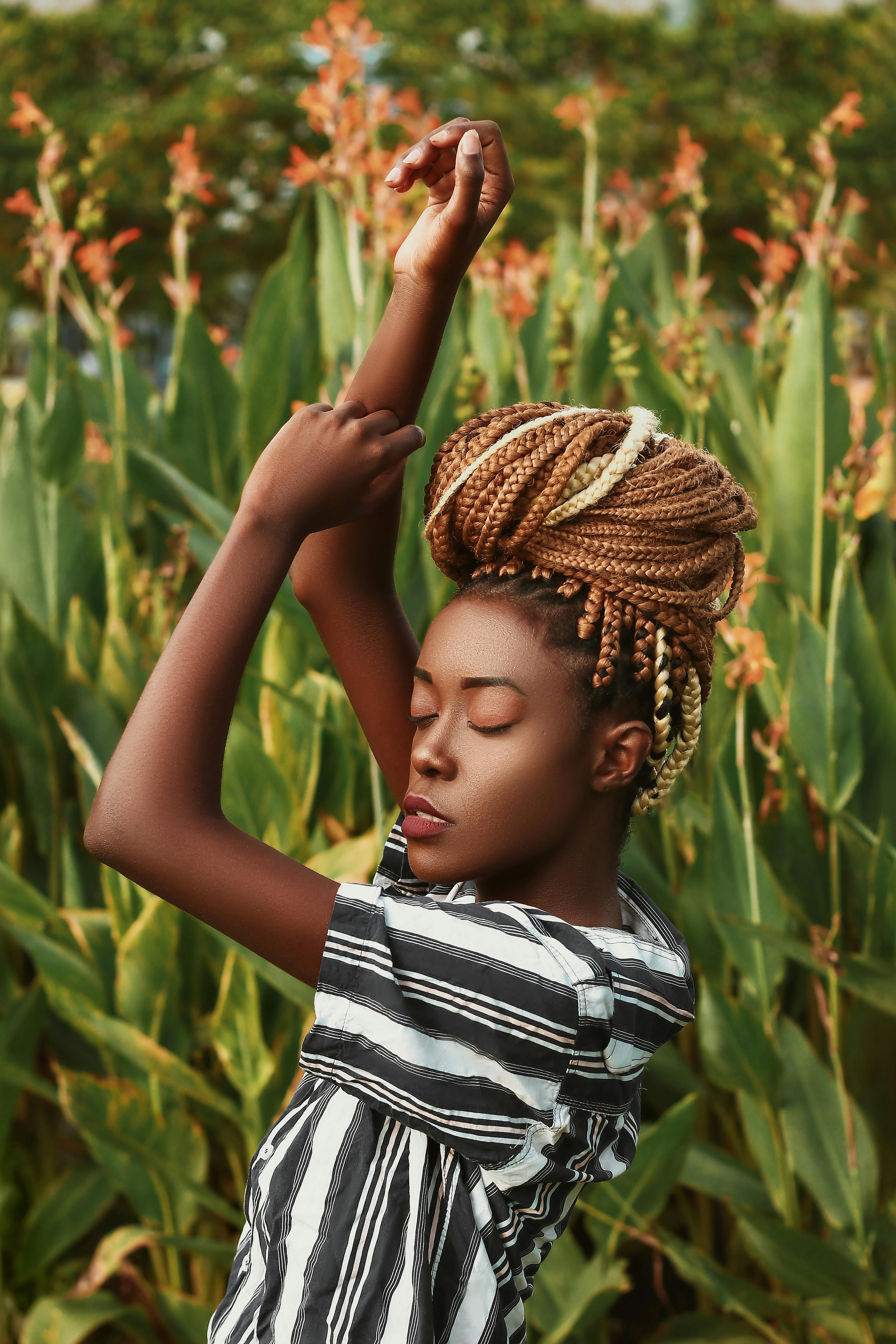 Side view of young African American female with braids in casual clothes standing with raised arms and closed eyes among green trees in countryside in sunny day