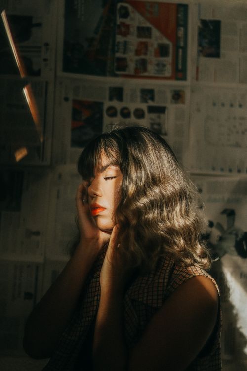 Free A woman with long hair and red lipstick poses thoughtfully in soft lighting against newspaper-covered walls. Stock Photo
