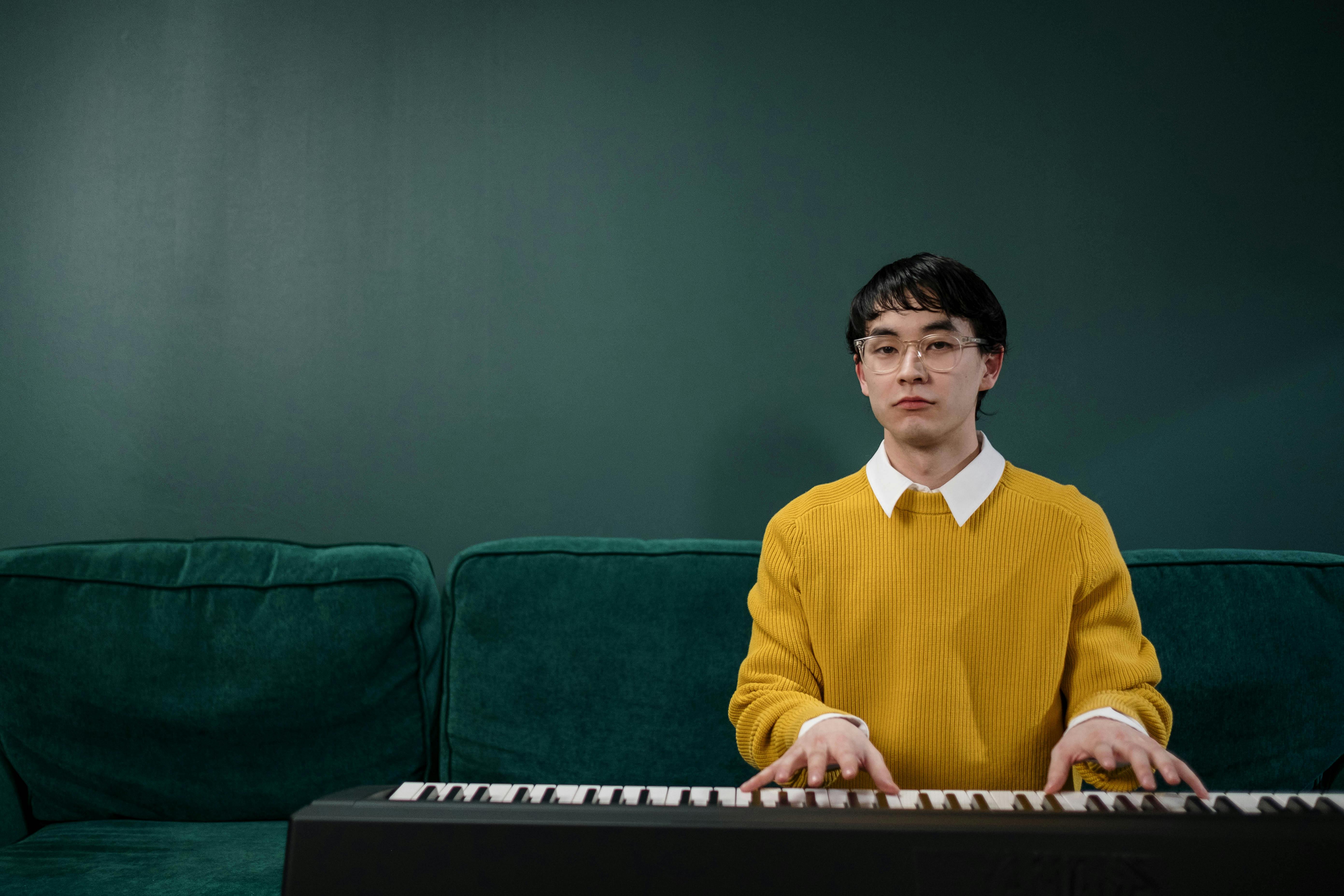 Young man in yellow sweater playing keyboard indoors with green backdrop.