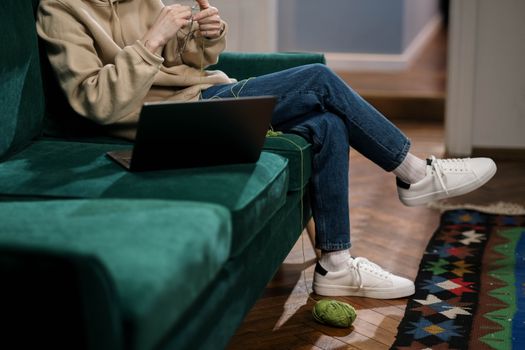 An adult knitting and using a laptop on a green sofa, showcasing a cozy indoor setup.