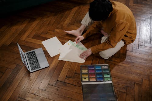 A Person Sitting on the Wooden Floor While Making a Drawing on the White Paper