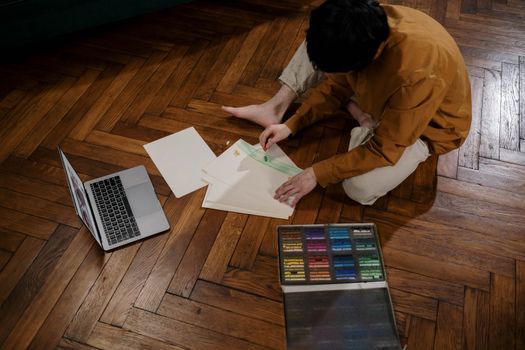 Person engaged in art activity on wooden floor with laptop and pastel colors.