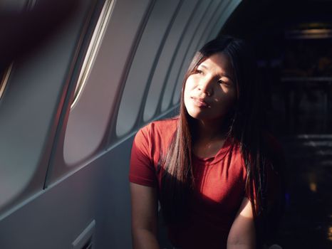 A woman in a red shirt sits in an airplane, gazing thoughtfully out the window as sunlight illuminates her face.