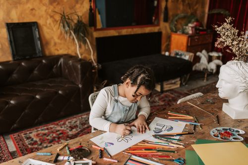 Free Young girl drawing at a table full of art supplies in a cozy living room setting. Stock Photo