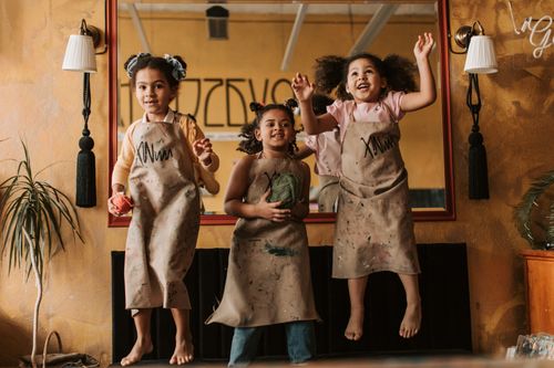 Free Three smiling girls wearing aprons jump joyfully indoors, reflected in a large mirror. Stock Photo