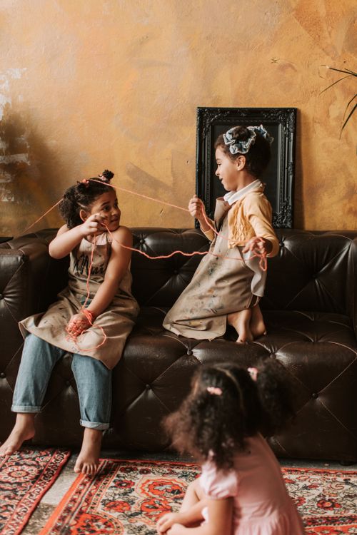 Free Three children joyfully play and interact with yarn on a sofa indoors, creating a playful and warm atmosphere. Stock Photo