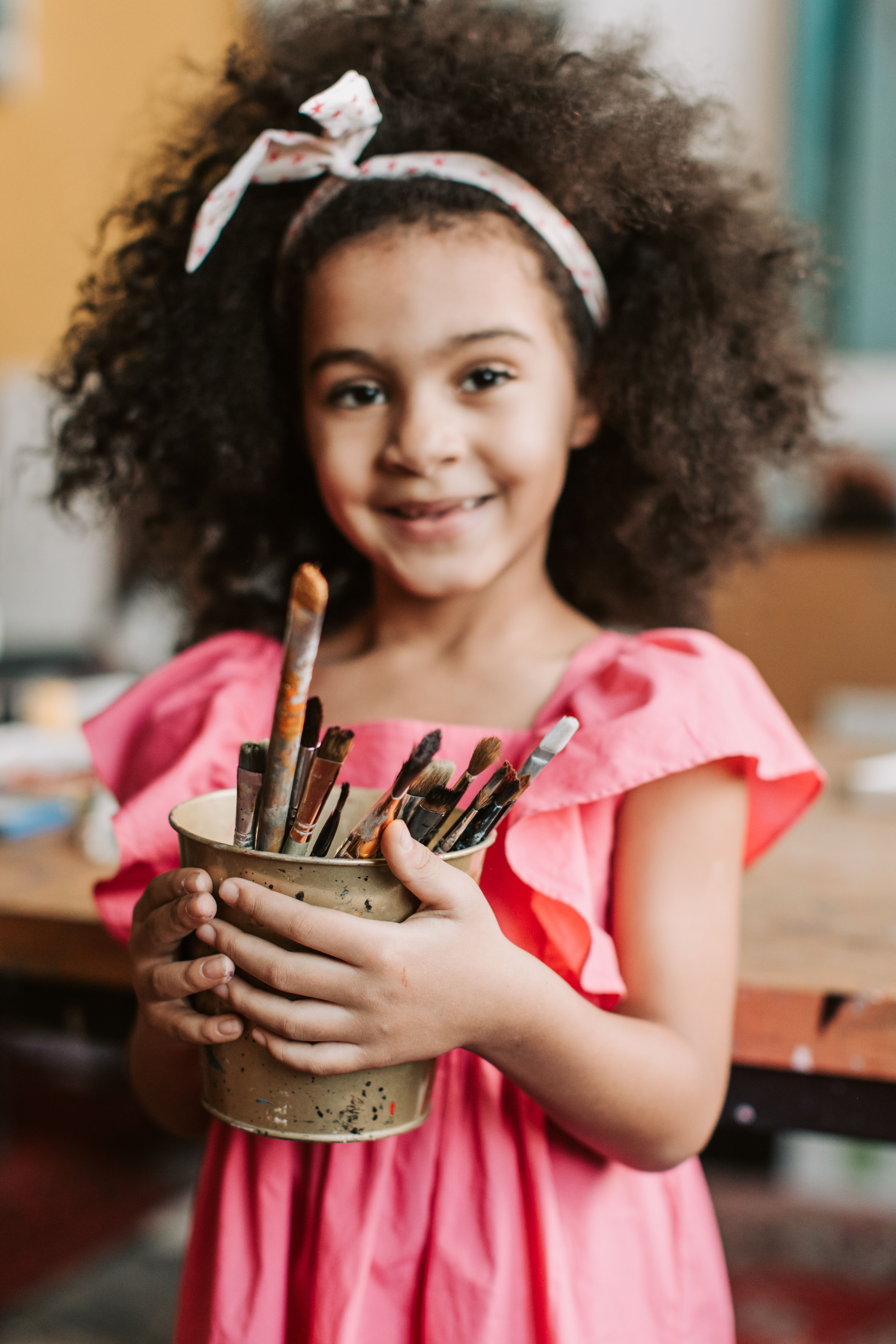 A happy girl holding a container of paintbrushes in an art studio, expressing creativity.