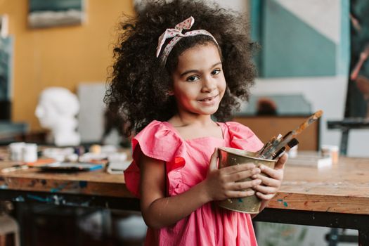 Adorable child with afro hair smiling and holding paint brushes indoors.