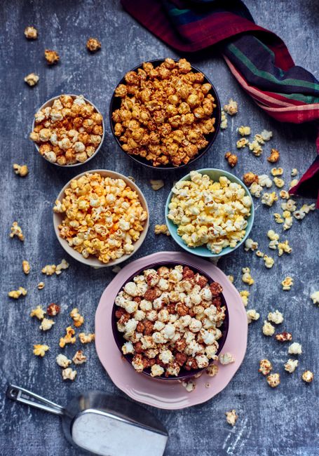 Top view of assorted flavored popcorn in various bowls on a rustic background with scattered kernels.