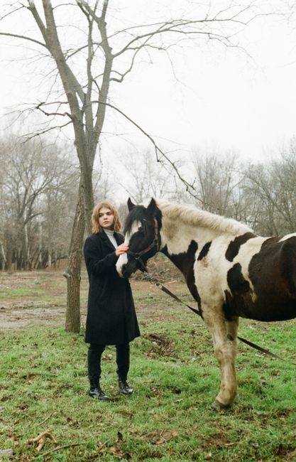A woman in black stands beside a spotted horse in a winter setting with bare trees.