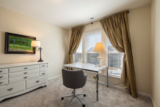 Cozy home office with glass desk, upholstered chair, and warm lighting by the window.