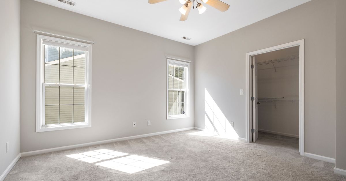 Sunlit empty room with beige carpet and ceiling fan, ideal for real estate listings.