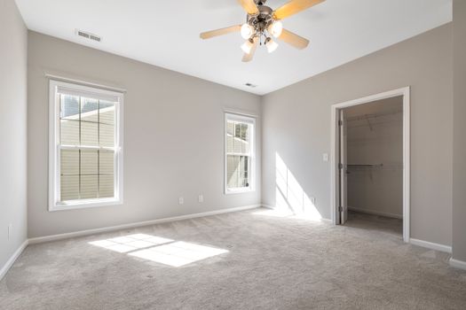 Sunlit empty room with beige carpet and ceiling fan, ideal for real estate listings.