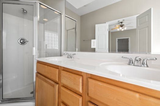 Bright and modern bathroom featuring a dual sink vanity, glass shower, and large mirror reflecting ceiling fan light.