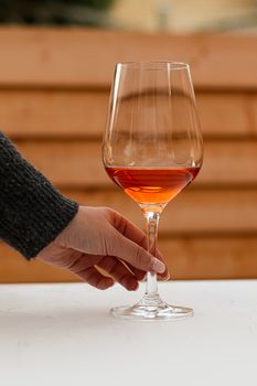 Hand holding a wine glass with an amber beverage outdoors against a wooden background.