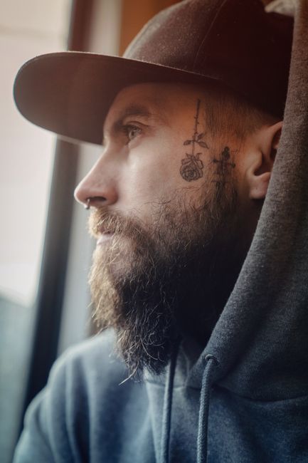 Profile of a bearded man with facial tattoos in a hoodie looking outside, creating a thoughtful mood.