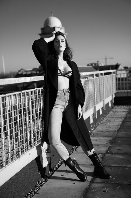 A confident woman poses in a black coat on a rooftop, exuding modern urban style.
