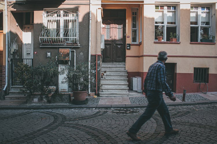 A Man Walking On A Road Near A House With A Cat On The Steps