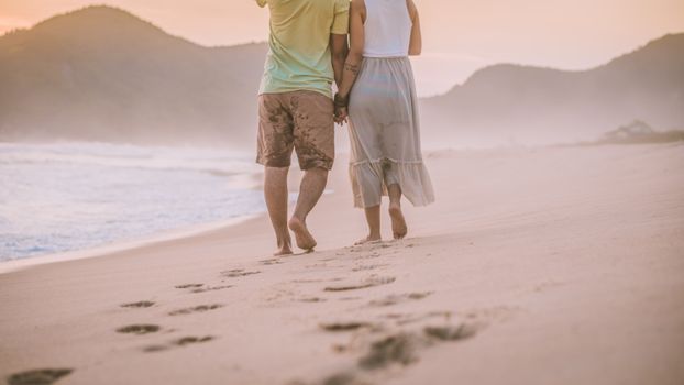 A couple holding hands and strolling on a serene beach with mountains in the background during sunrise.