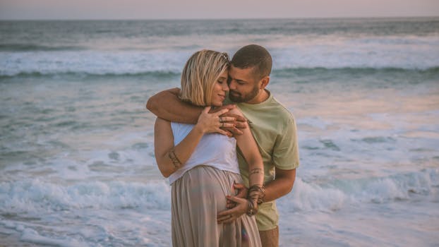 Affectionate couple embracing on a serene beach at sunset, celebrating pregnancy.