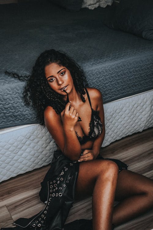 Free A Woman Wearing Black Brassiere Sitting on a Wooden Floor Beside a Bed Stock Photo