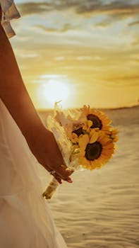 A person holds a bouquet of sunflowers against a glowing sunset by the beach.