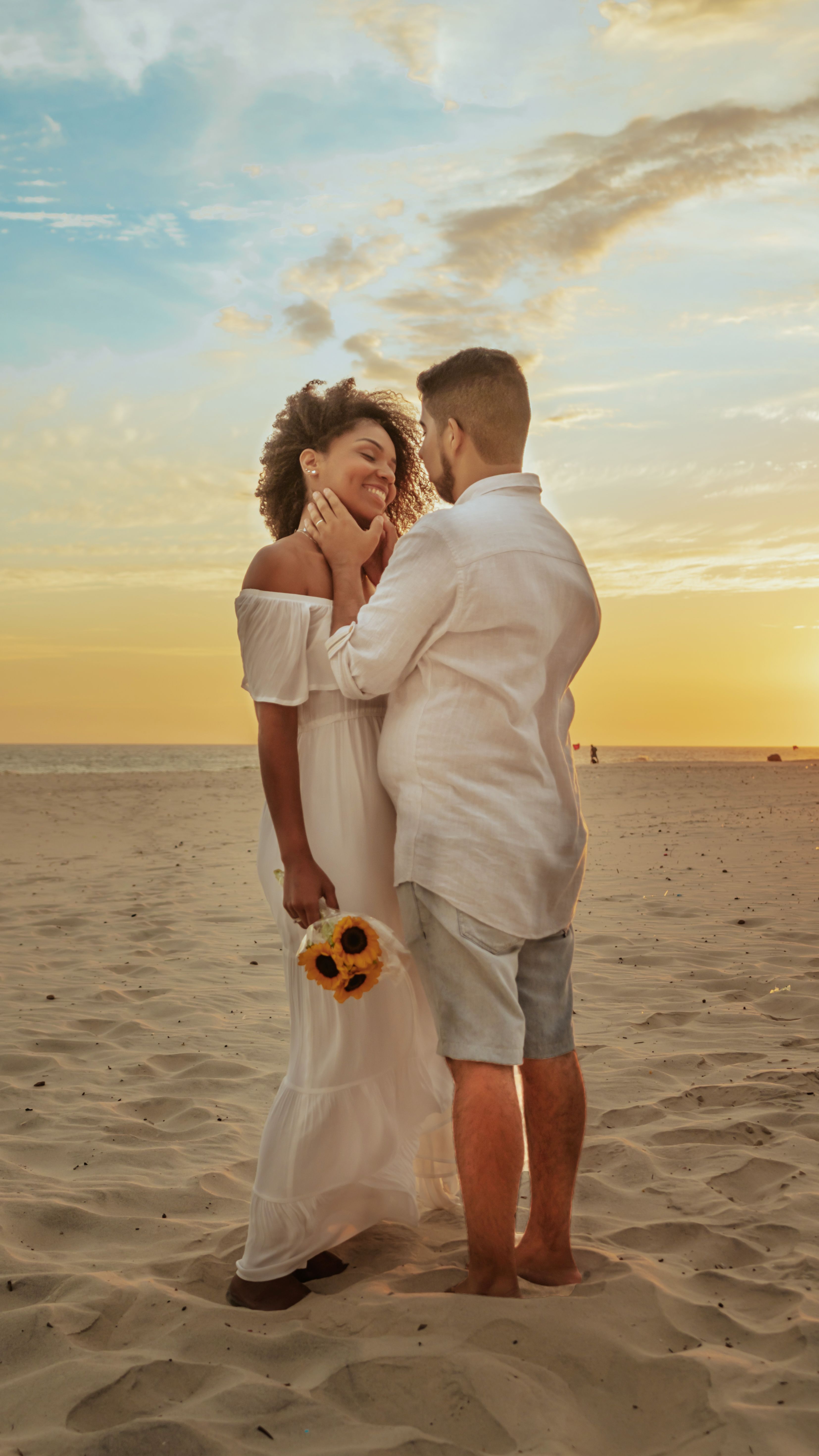 A couple in white attire sharing a romantic moment on a sandy beach during a beautiful sunset.