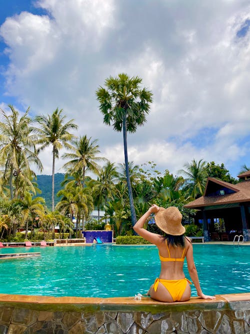 A Woman in Yellow Bikini Sitting on the Poolside while Wearing a Hat