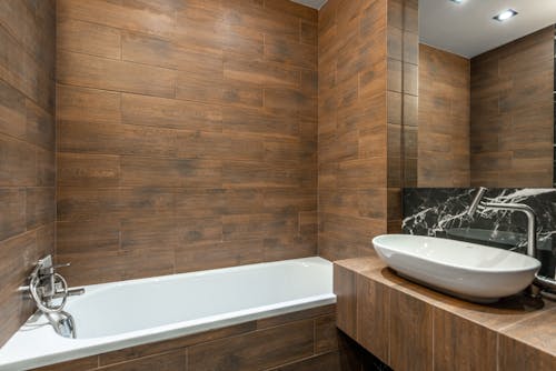 Stylish interior design of modern bathroom with wood like tiles equipped with white bathtub and sink with faucet
