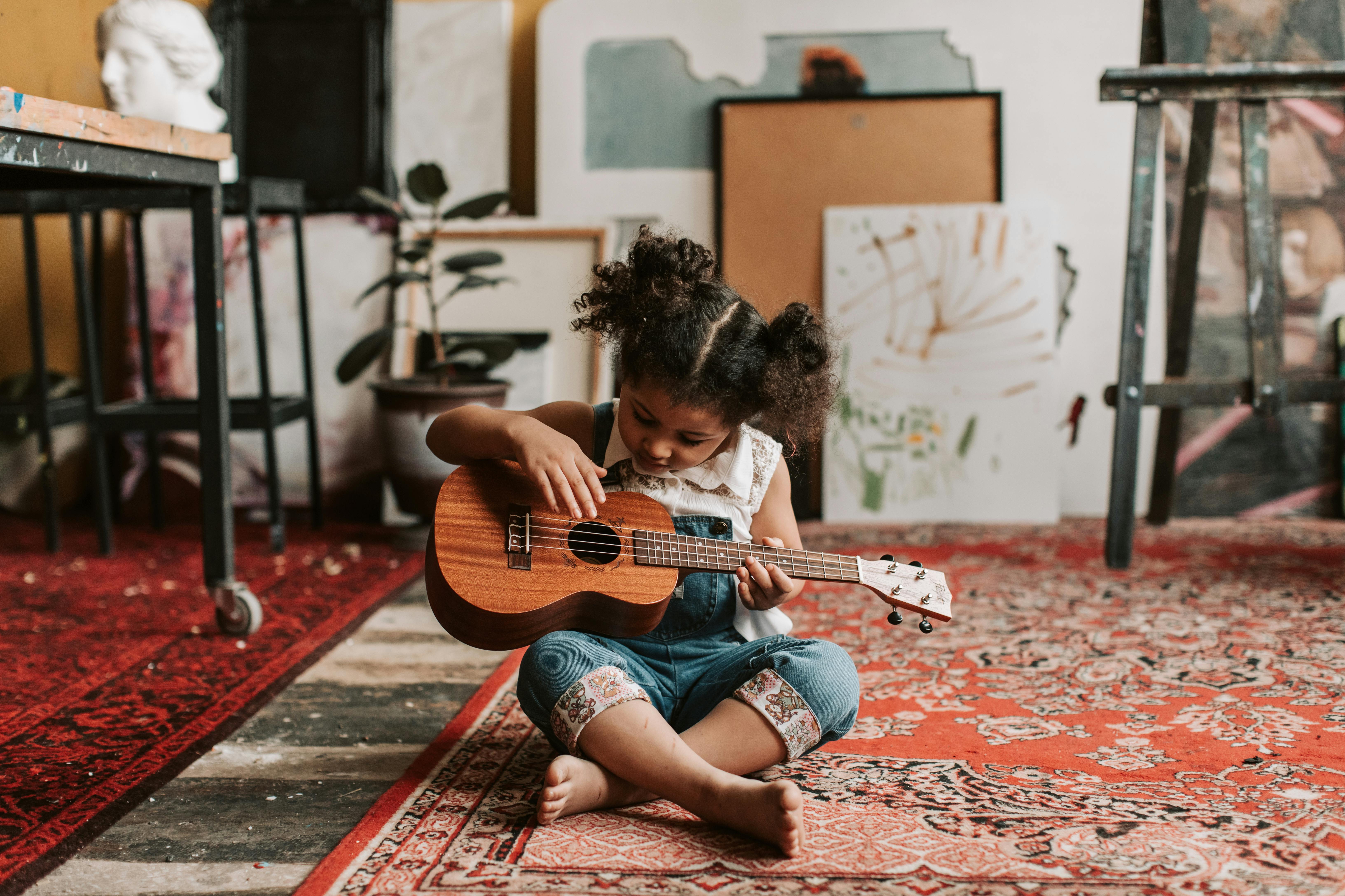 a young girl in denim jumper sitting on the floor while playing ukulele