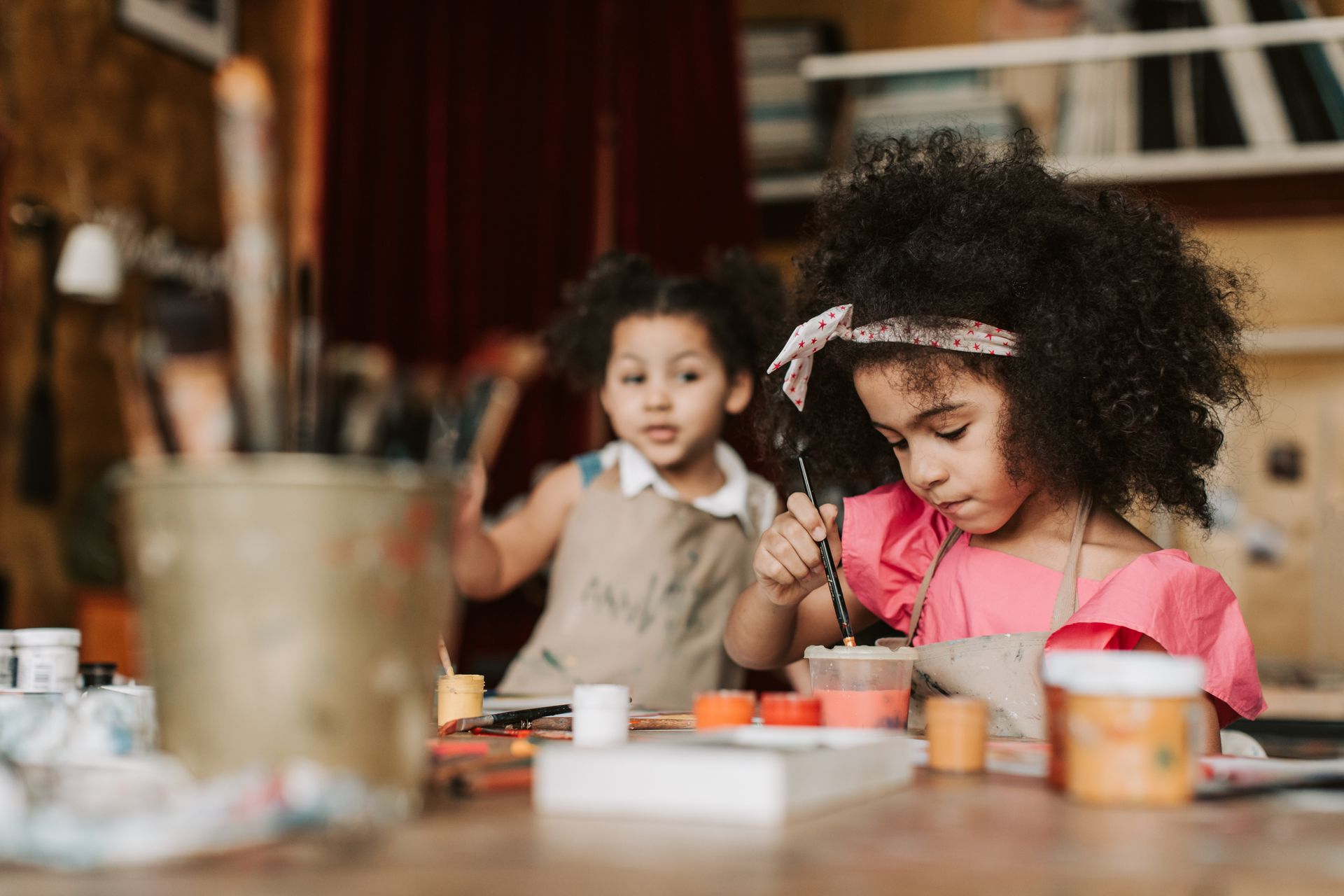 Two young girls engaged in painting activities in a cozy art workshop with natural light and warm tones.