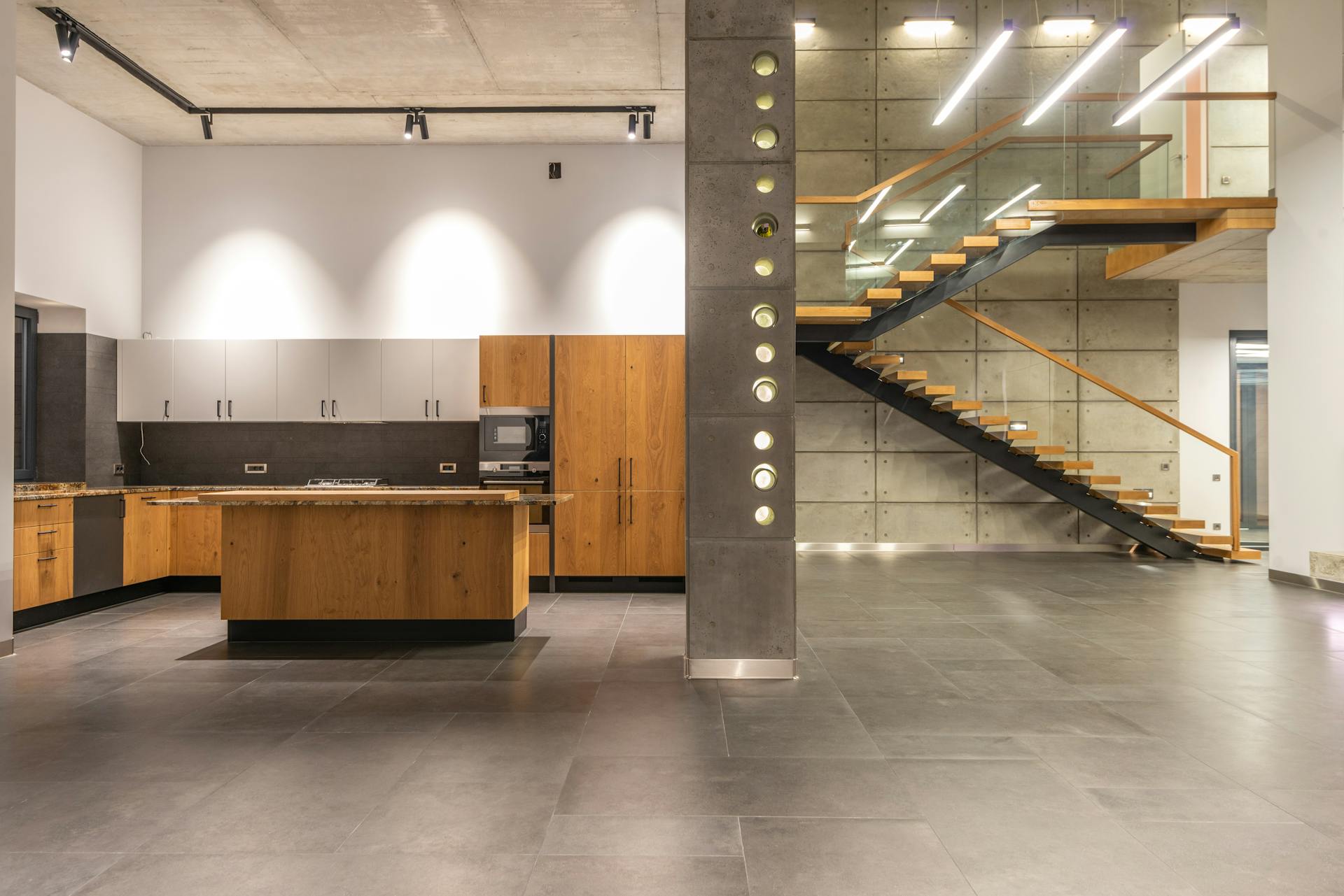 Interior design of spacious open plan kitchen with wooden furniture and hallway with stairs leading to upper storey in contemporary loft style house with stony tiles and LED lights