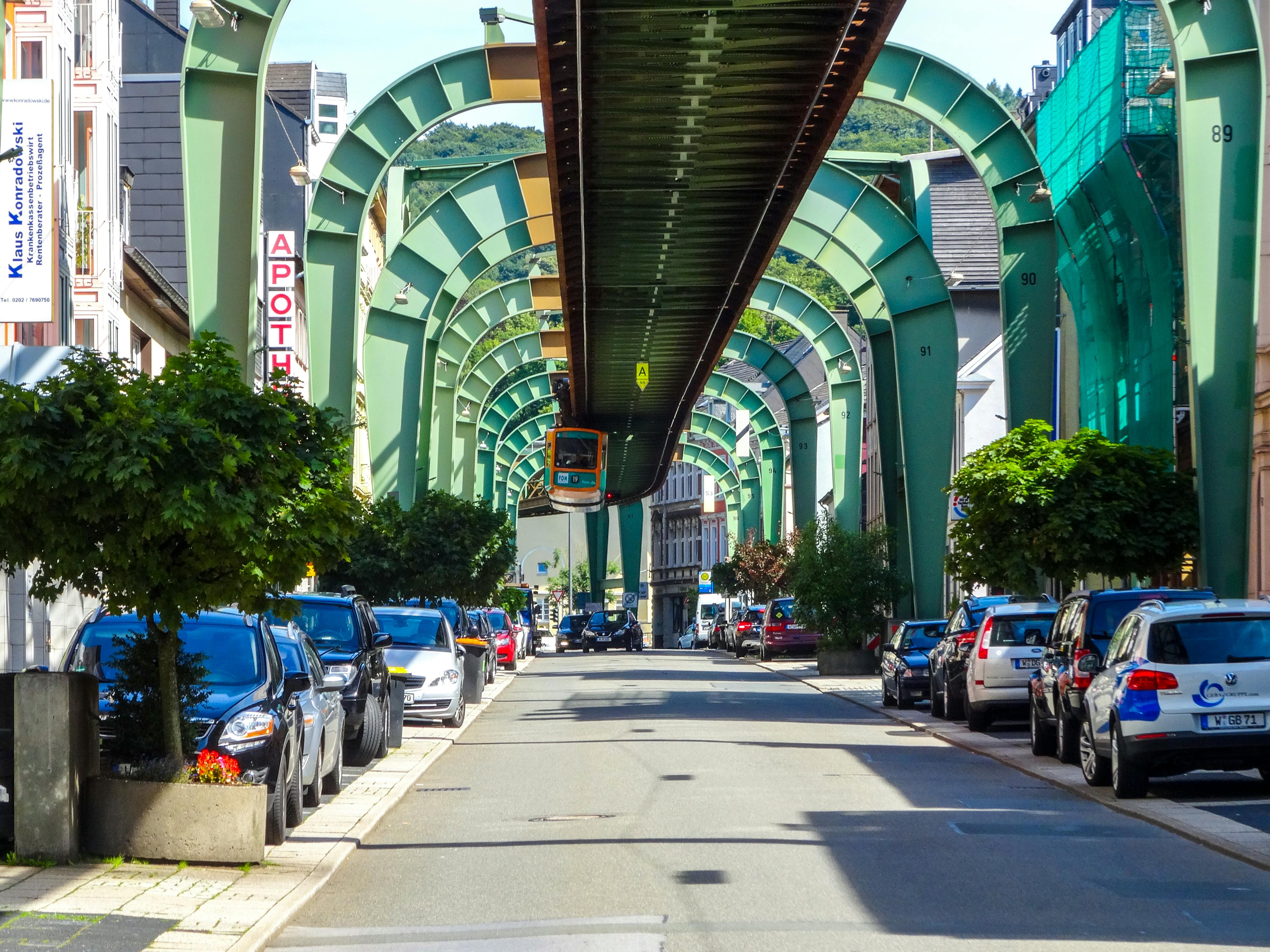 Wuppertal's suspension railway travels above a street lined with parked cars and buildings.