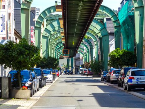 Wuppertal's suspension railway travels above a street lined with parked cars and buildings.