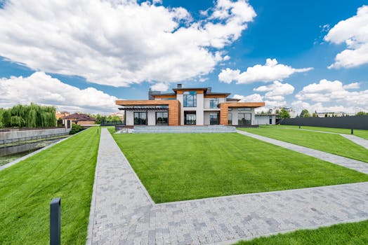 Picturesque landscape of modern house with lawn and cobblestone paths against cloudy blue sky and trees on background