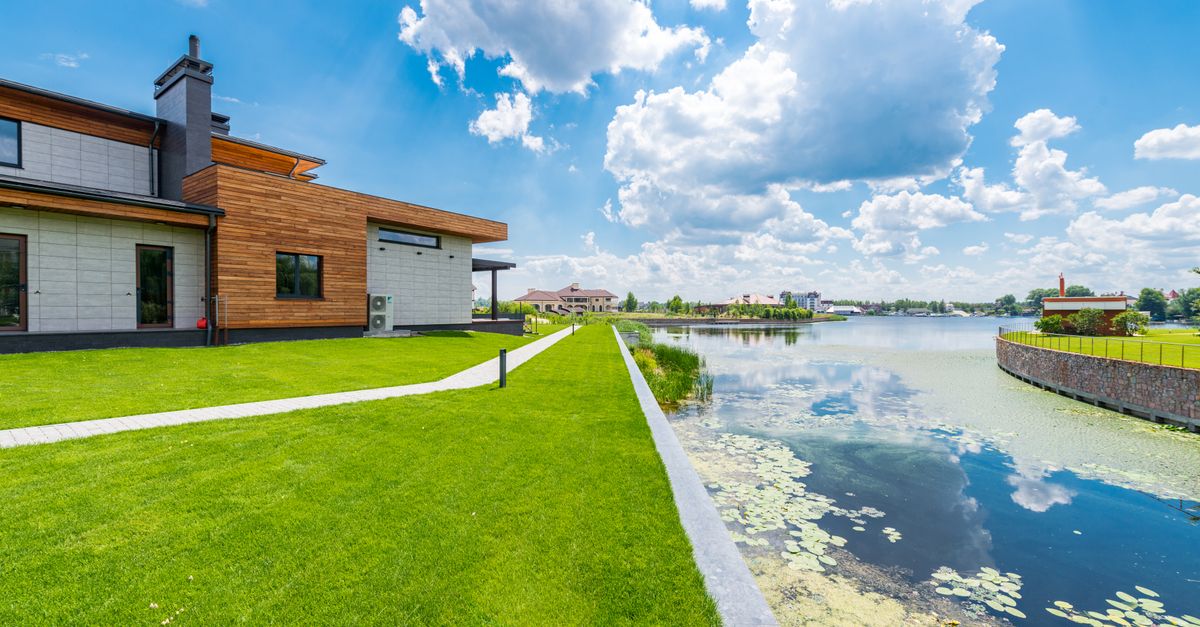 Exterior of contemporary residential cottage with windows and glass doors located on green lawn near pond against blue sky with clouds on sunny day in summer