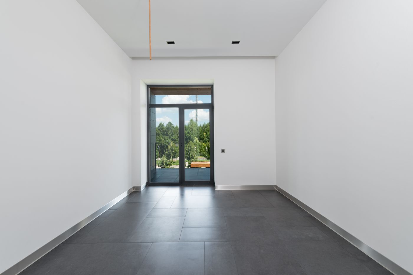 An empty finished room with large dark grey floor tiles, white walls, skirting boards and glass doors opening outdoors.