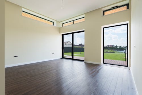 Interior of spacious room with wooden laminate floor big windows and glass door viewing terrace and green lawn