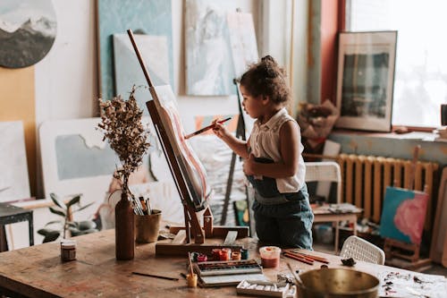 A Young Girl Using a Paintbrush while Standing Near the Wooden Table